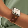 Slim vegan leather Apple Watch band with brown linen texture, featuring detailed embroidery of red cherries, pink bows, and wildflowers. Shows silver metal buckle and contrasting olive green leather loops. Perfect gift for her.
