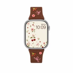 Slim vegan leather Apple Watch band with brown linen texture, featuring detailed embroidery of red cherries, pink bows, and wildflowers. Shows silver metal buckle and contrasting olive green leather loops. Perfect gift for her.