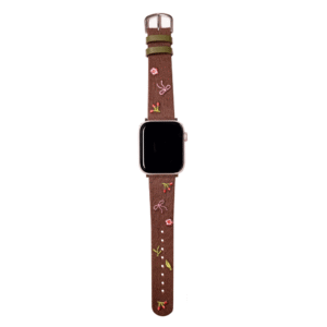 Slim vegan leather Apple Watch band with brown linen texture, featuring detailed embroidery of red cherries, pink bows, and wildflowers. Shows silver metal buckle and contrasting olive green leather loops. Perfect gift for her.