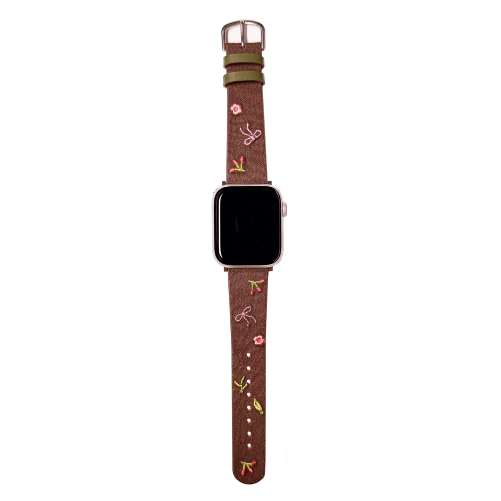 Slim vegan leather Apple Watch band with brown linen texture, featuring detailed embroidery of red cherries, pink bows, and wildflowers. Shows silver metal buckle and contrasting olive green leather loops. Perfect gift for her.