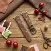 Slim vegan leather Apple Watch band with brown linen texture, featuring detailed embroidery of red cherries, pink bows, and wildflowers. Shows silver metal buckle and contrasting olive green leather loops. Perfect gift for her.