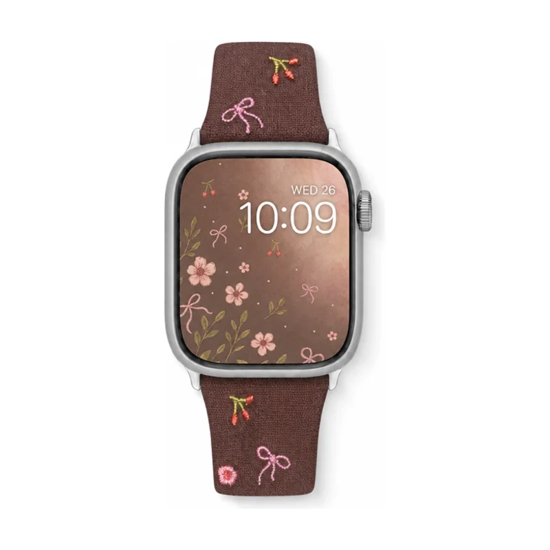 Slim vegan leather Apple Watch band with brown linen texture, featuring detailed embroidery of red cherries, pink bows, and wildflowers. Shows silver metal buckle and contrasting olive green leather loops. Perfect gift for her.