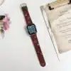 Slim vegan leather Apple Watch band with brown linen texture, featuring detailed embroidery of red cherries, pink bows, and wildflowers. Shows silver metal buckle and contrasting olive green leather loops. Perfect gift for her.