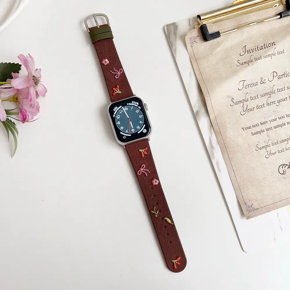 Slim vegan leather Apple Watch band with brown linen texture, featuring detailed embroidery of red cherries, pink bows, and wildflowers. Shows silver metal buckle and contrasting olive green leather loops. Perfect gift for her.