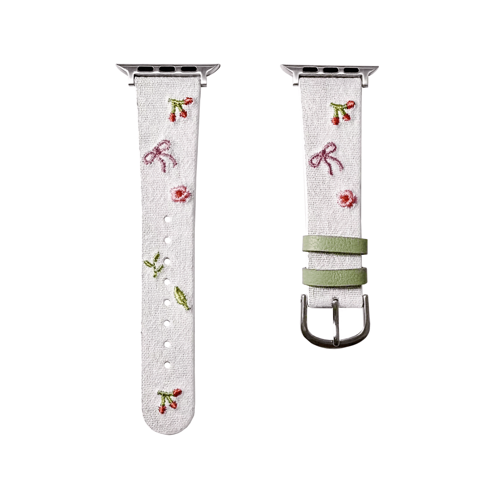 Slim vegan leather Apple Watch band with white linen texture, featuring detailed embroidery of red cherries, pink bows, and wildflowers. Shows silver metal buckle and contrasting olive green leather loops. Perfect gift for her.