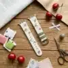 Slim vegan leather Apple Watch band with white linen texture, featuring detailed embroidery of red cherries, pink bows, and wildflowers. Shows silver metal buckle and contrasting olive green leather loops. Perfect gift for her.