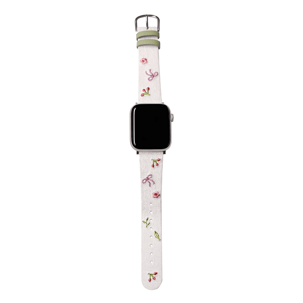 Slim vegan leather Apple Watch band with white linen texture, featuring detailed embroidery of red cherries, pink bows, and wildflowers. Shows silver metal buckle and contrasting olive green leather loops. Perfect gift for her.