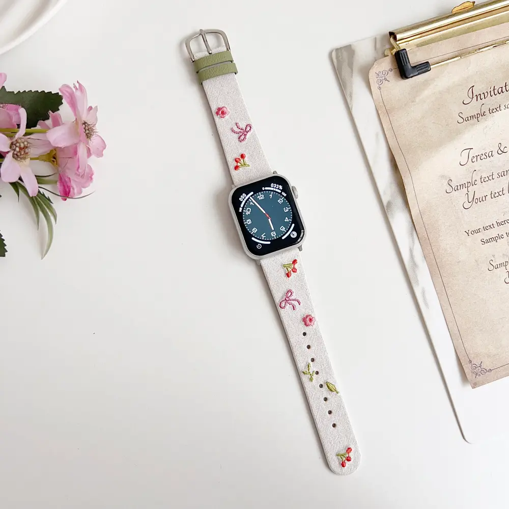 Slim vegan leather Apple Watch band with white linen texture, featuring detailed embroidery of red cherries, pink bows, and wildflowers. Shows silver metal buckle and contrasting olive green leather loops. Perfect gift for her.
