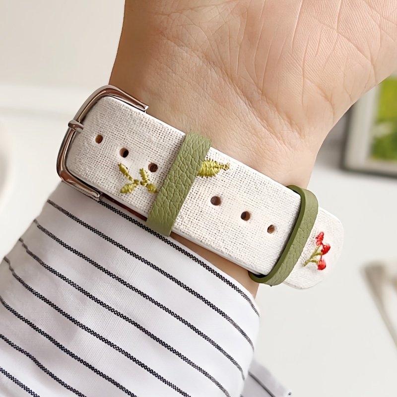 Slim vegan leather Apple Watch band with white linen texture, featuring detailed embroidery of red cherries, pink bows, and wildflowers. Shows silver metal buckle and contrasting olive green leather loops. Perfect gift for her.