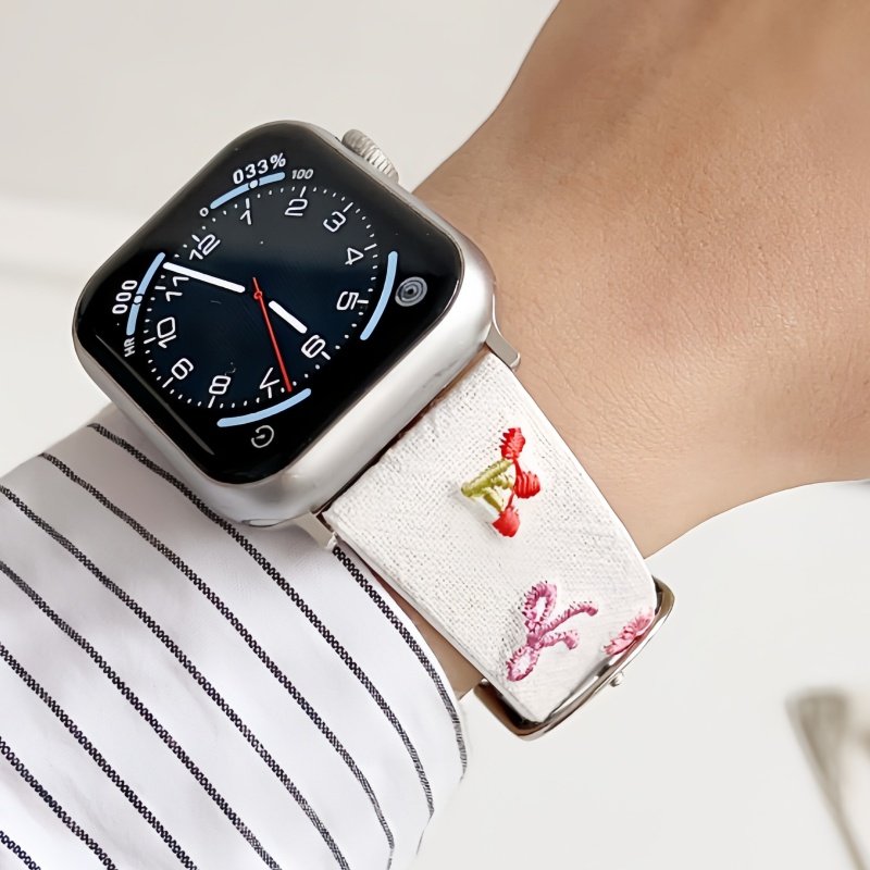 Slim vegan leather Apple Watch band with white linen texture, featuring detailed embroidery of red cherries, pink bows, and wildflowers. Shows silver metal buckle and contrasting olive green leather loops. Perfect gift for her.