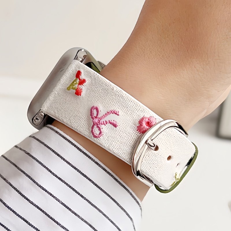 Slim vegan leather Apple Watch band with white linen texture, featuring detailed embroidery of red cherries, pink bows, and wildflowers. Shows silver metal buckle and contrasting olive green leather loops. Perfect gift for her.