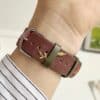 Slim vegan leather Apple Watch band with brown linen texture, featuring detailed embroidery of red cherries, pink bows, and wildflowers. Shows silver metal buckle and contrasting olive green leather loops. Perfect gift for her.