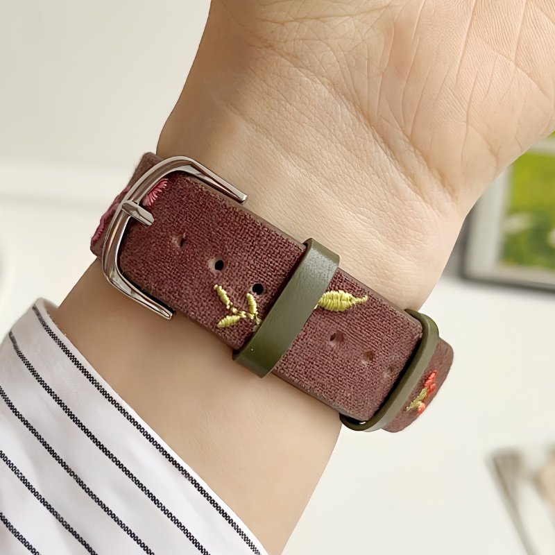 Slim vegan leather Apple Watch band with brown linen texture, featuring detailed embroidery of red cherries, pink bows, and wildflowers. Shows silver metal buckle and contrasting olive green leather loops. Perfect gift for her.