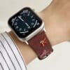 Slim vegan leather Apple Watch band with brown linen texture, featuring detailed embroidery of red cherries, pink bows, and wildflowers. Shows silver metal buckle and contrasting olive green leather loops. Perfect gift for her.