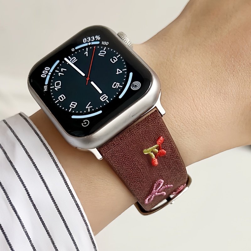 Slim vegan leather Apple Watch band with brown linen texture, featuring detailed embroidery of red cherries, pink bows, and wildflowers. Shows silver metal buckle and contrasting olive green leather loops. Perfect gift for her.