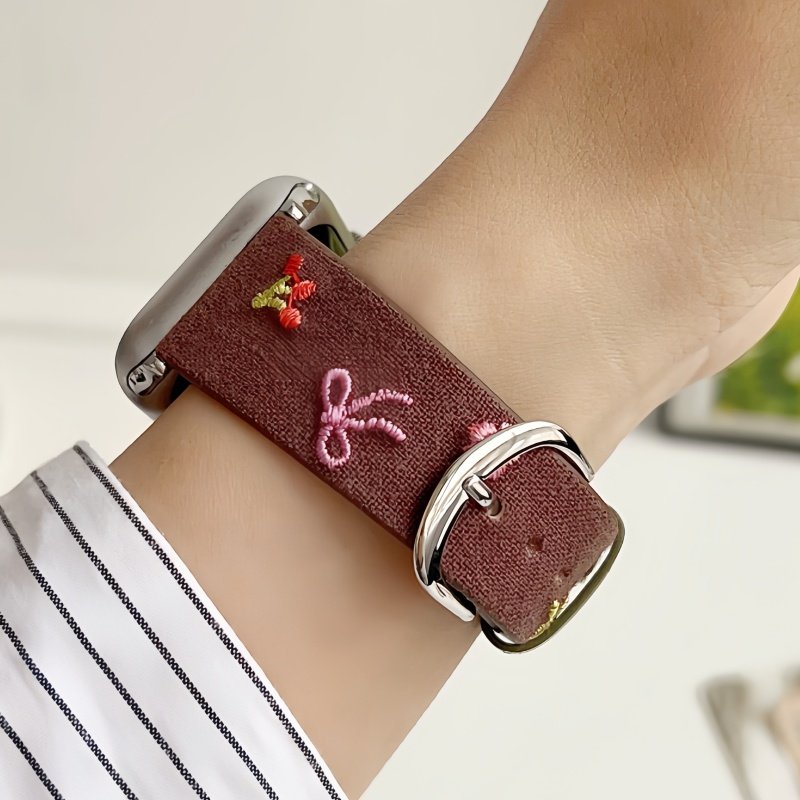 Slim vegan leather Apple Watch band with brown linen texture, featuring detailed embroidery of red cherries, pink bows, and wildflowers. Shows silver metal buckle and contrasting olive green leather loops. Perfect gift for her.