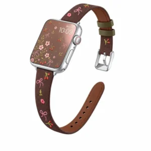 Slim vegan leather Apple Watch band with brown linen texture, featuring detailed embroidery of red cherries, pink bows, and wildflowers. Shows silver metal buckle and contrasting olive green leather loops. Perfect gift for her.