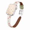 Slim vegan leather Apple Watch band with white linen texture, featuring detailed embroidery of red cherries, pink bows, and wildflowers. Shows silver metal buckle and contrasting olive green leather loops. Perfect gift for her.