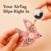 Hand holding an Apple AirTag beside a coral pink vegan leather bird AirTag holder with gold clip.