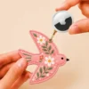 Hand holding an Apple AirTag beside a coral pink vegan leather bird AirTag holder with gold clip.