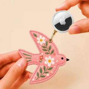 Hand holding an Apple AirTag beside a coral pink vegan leather bird AirTag holder with gold clip.