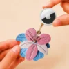 Hand holding an Apple AirTag beside a pink and blue vegan leather lily flower AirTag holder with gold clip