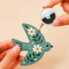 Hand holding an Apple AirTag beside a sage green vegan leather bird AirTag holder with gold clip.