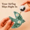 Hand holding an Apple AirTag beside a sage green vegan leather bird AirTag holder with gold clip.