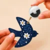 Hand holding an Apple AirTag beside a navy blue vegan leather bird AirTag holder with gold clip.