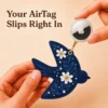 Hand holding an Apple AirTag beside a navy blue vegan leather bird AirTag holder with gold clip.