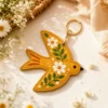 Mustard yellow vegan leather AirTag case shaped like a bird with white daisies and gold keyring clip