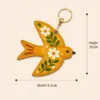 Mustard yellow vegan leather AirTag case shaped like a bird with white daisies and gold keyring clip
