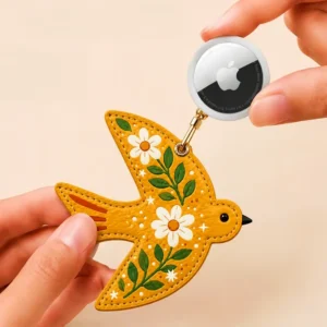 Mustard yellow vegan leather AirTag case shaped like a bird with white daisies and gold keyring clip